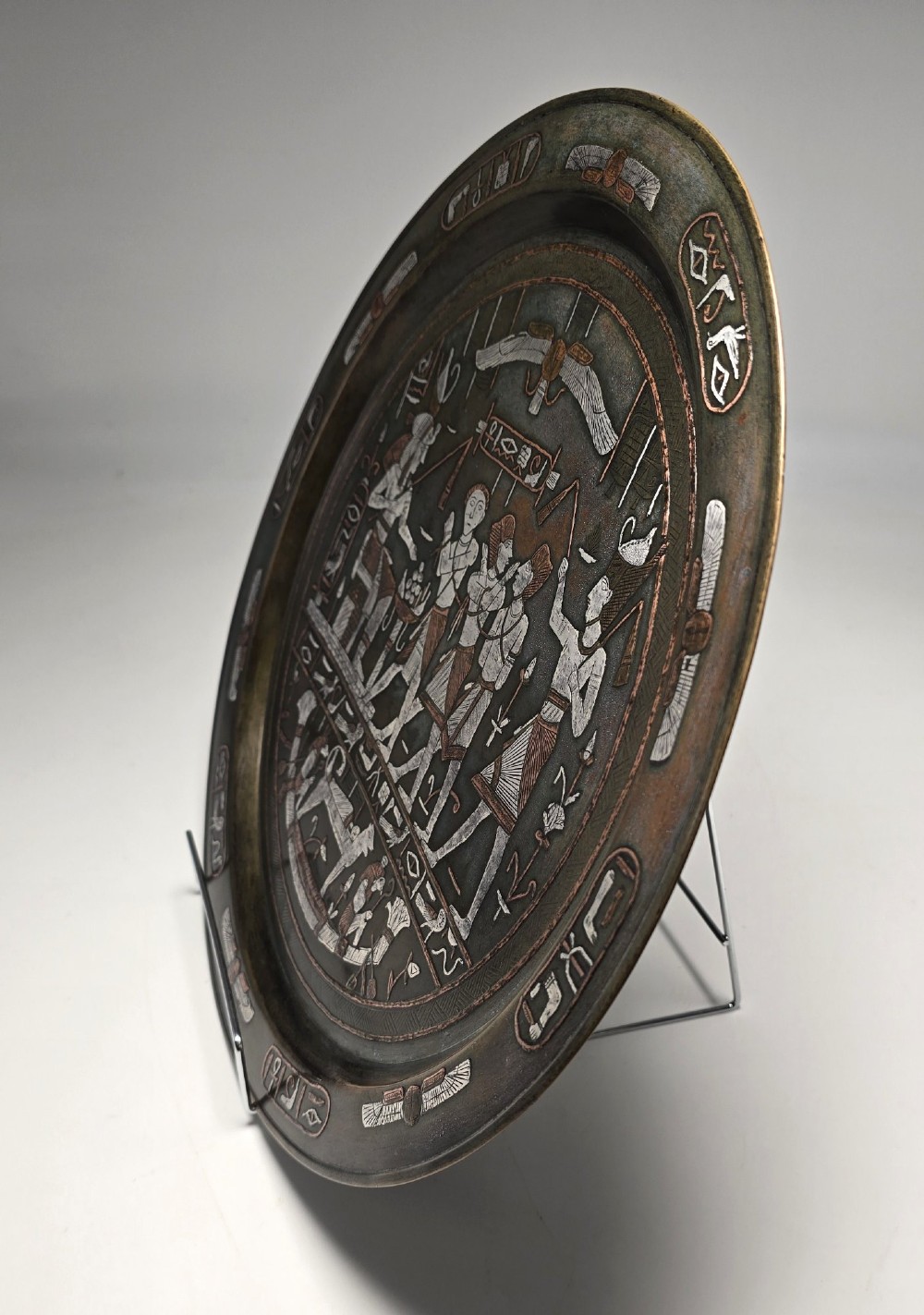 A circular bronze charger decorated with silver and copper inlays depicting pharaonic figures, hieroglyphs, and a winged sun deity. View XI