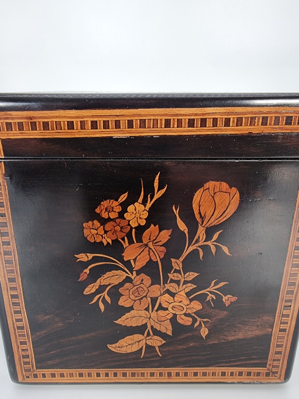 A square ebonised wooden box featuring detailed Sorrento marquetry inlay depicting figures on the top lid, floral motifs on the sides, and geometric borders. View XV