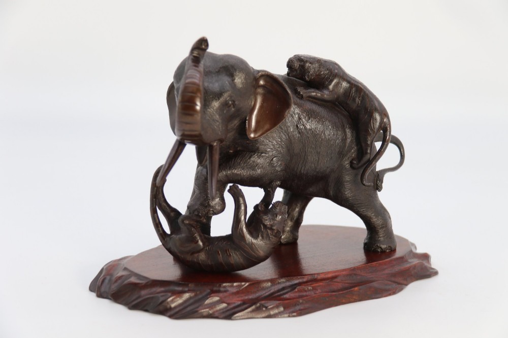 A dark patinated bronze sculpture of an elephant defending itself against two tigers, mounted on a carved wooden base. View II