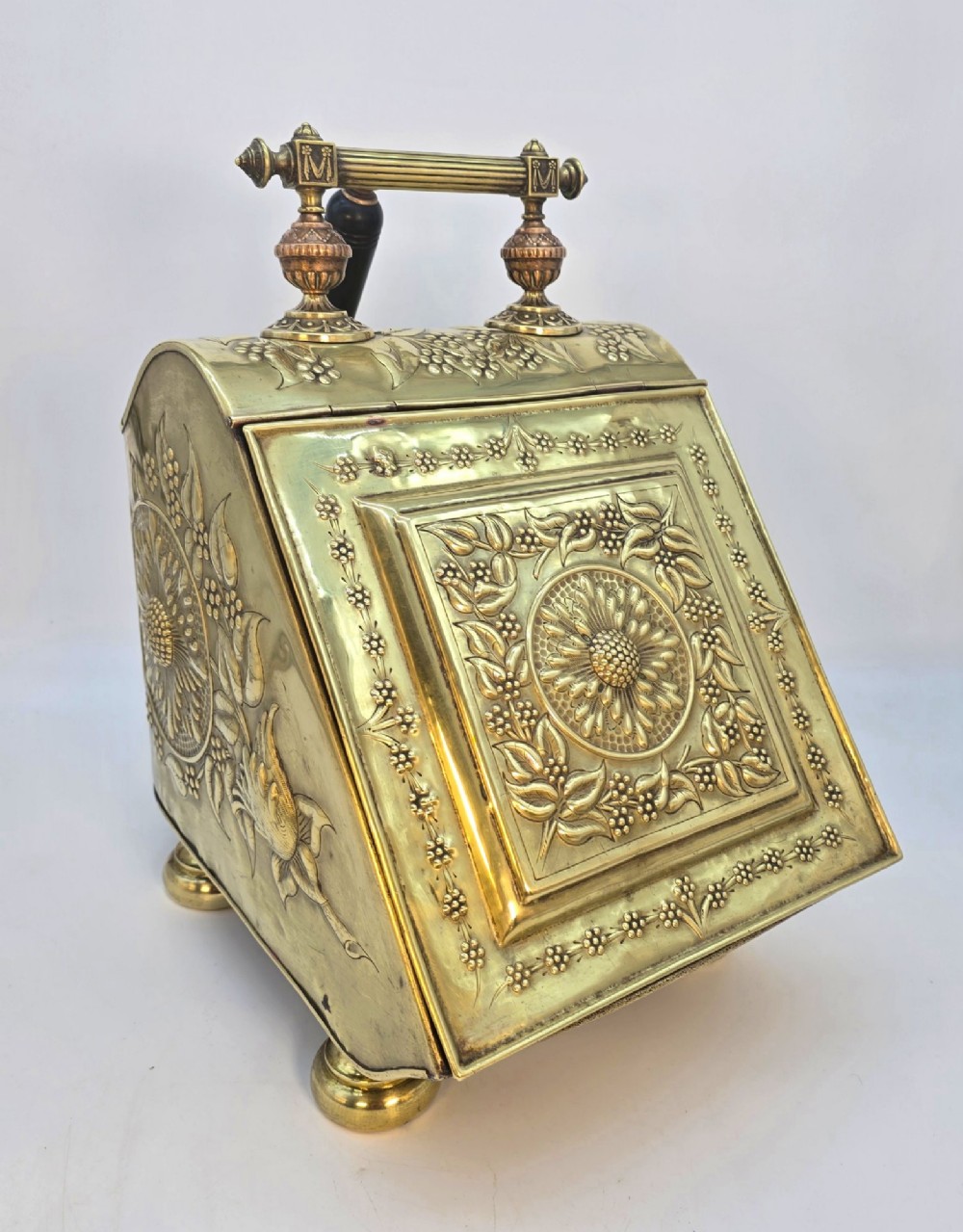Victorian sloped brass coal box featuring repousse sunflower motifs, turned brass feet, and a top carrying handle with an integrated rear shovel.