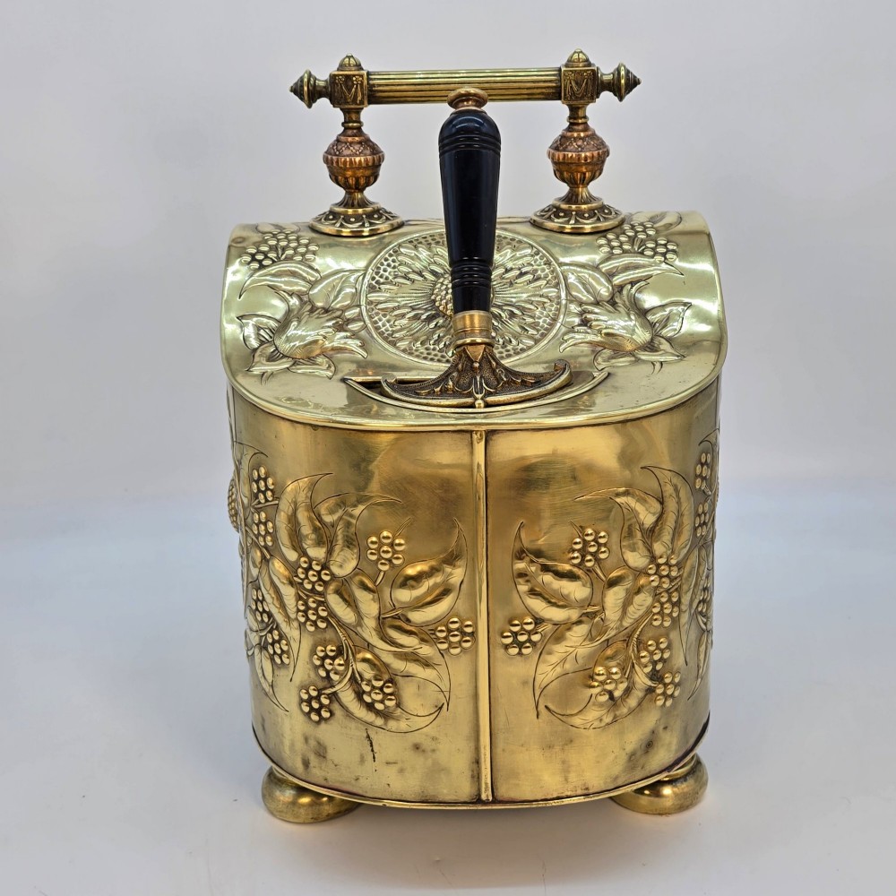 Victorian sloped brass coal box featuring repousse sunflower motifs, turned brass feet, and a top carrying handle with an integrated rear shovel. View VI