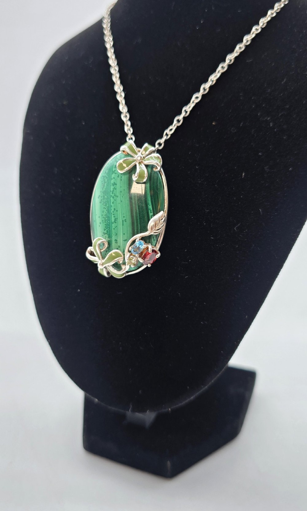 An oval green malachite pendant framed in silver metalwork with enamelled floral motifs, attached to a silver chain. View IX