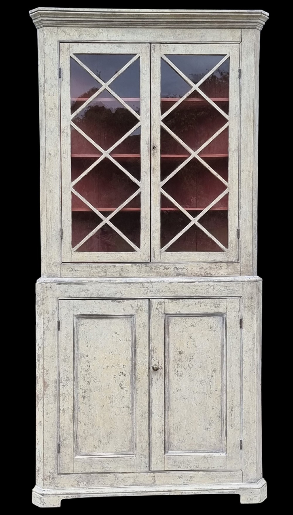 An antique two-part painted corner cupboard featuring an upper section with diamond-latticed glass doors showing a red painted interior and a solid two-door lower cabinet.