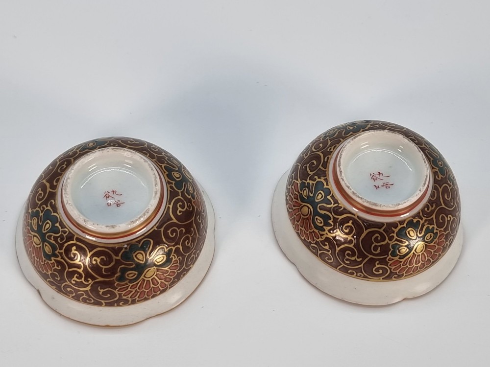 A pair of small, round Japanese porcelain bowls featuring intricate floral painted interiors and detailed red and gold geometric borders around scalloped rims. View IX