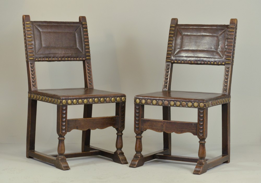 A pair of Victorian Spanish Revival oak dining chairs featuring brown leather seats, embossed back panels, and large brass stud detailing.