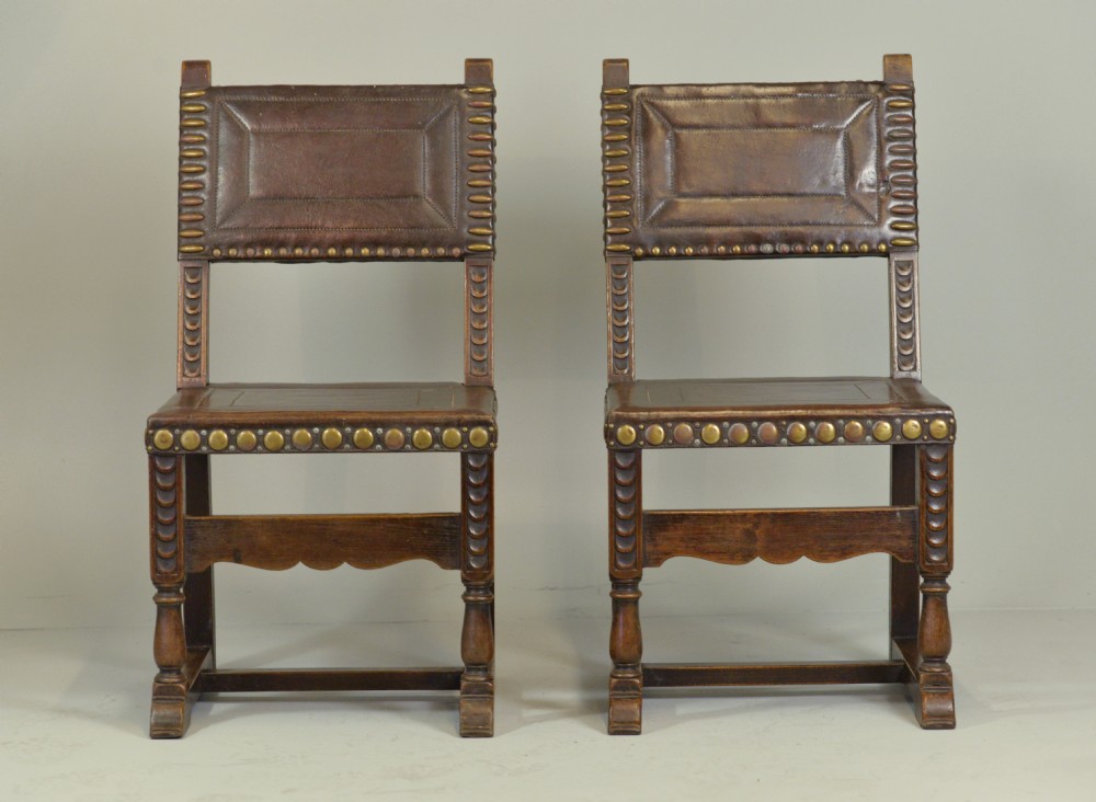 A pair of Victorian Spanish Revival oak dining chairs featuring brown leather seats, embossed back panels, and large brass stud detailing. View II
