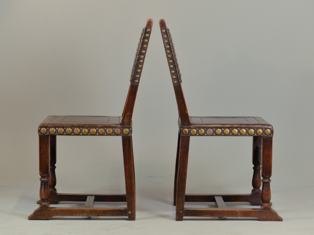 A pair of Victorian Spanish Revival oak dining chairs featuring brown leather seats, embossed back panels, and large brass stud detailing. View III