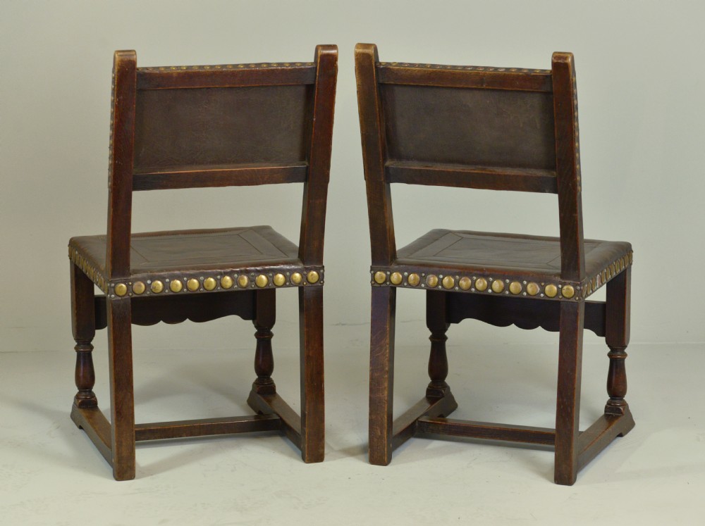 A pair of Victorian Spanish Revival oak dining chairs featuring brown leather seats, embossed back panels, and large brass stud detailing. View IV