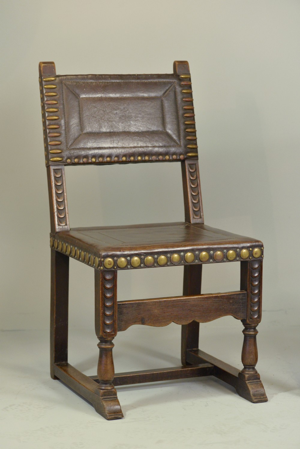 A pair of Victorian Spanish Revival oak dining chairs featuring brown leather seats, embossed back panels, and large brass stud detailing. View V