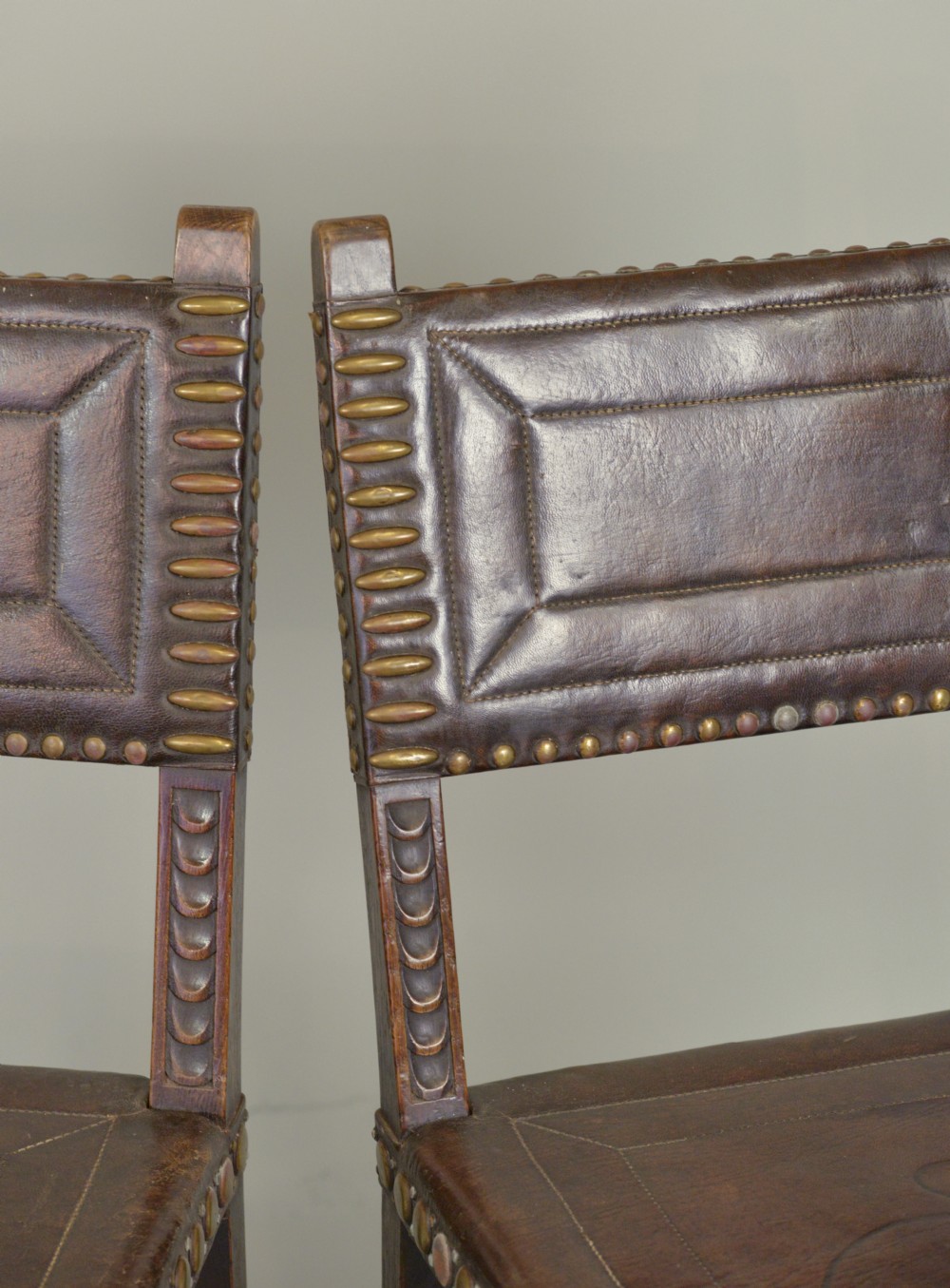 A pair of Victorian Spanish Revival oak dining chairs featuring brown leather seats, embossed back panels, and large brass stud detailing. View VII