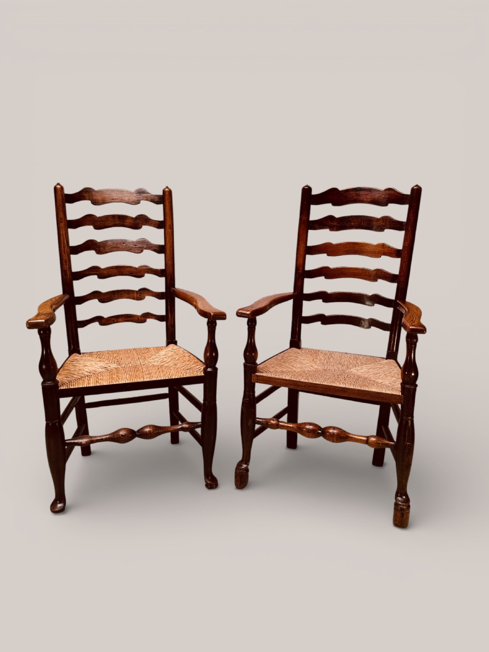 A pair of dark ash carver dining chairs featuring shaped wavy ladderback slats, turned front legs, and woven rush seats.