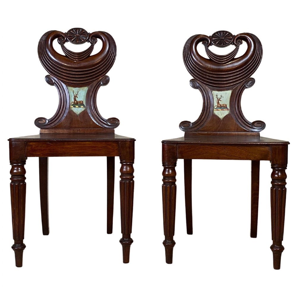 A pair of carved dark mahogany Georgian hall chairs featuring scroll-topped backs with painted stag crests and reeded turned front legs.