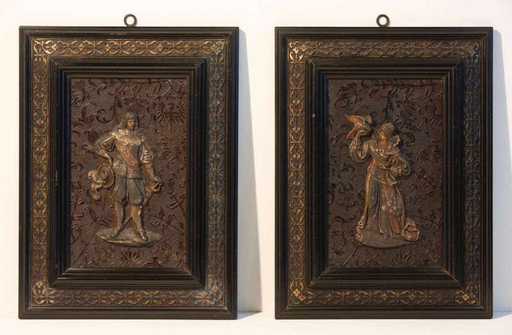 A pair of rectangular dark framed wall plaques displaying cast metal relief figures of a seventeenth-century cavalier and falconer.