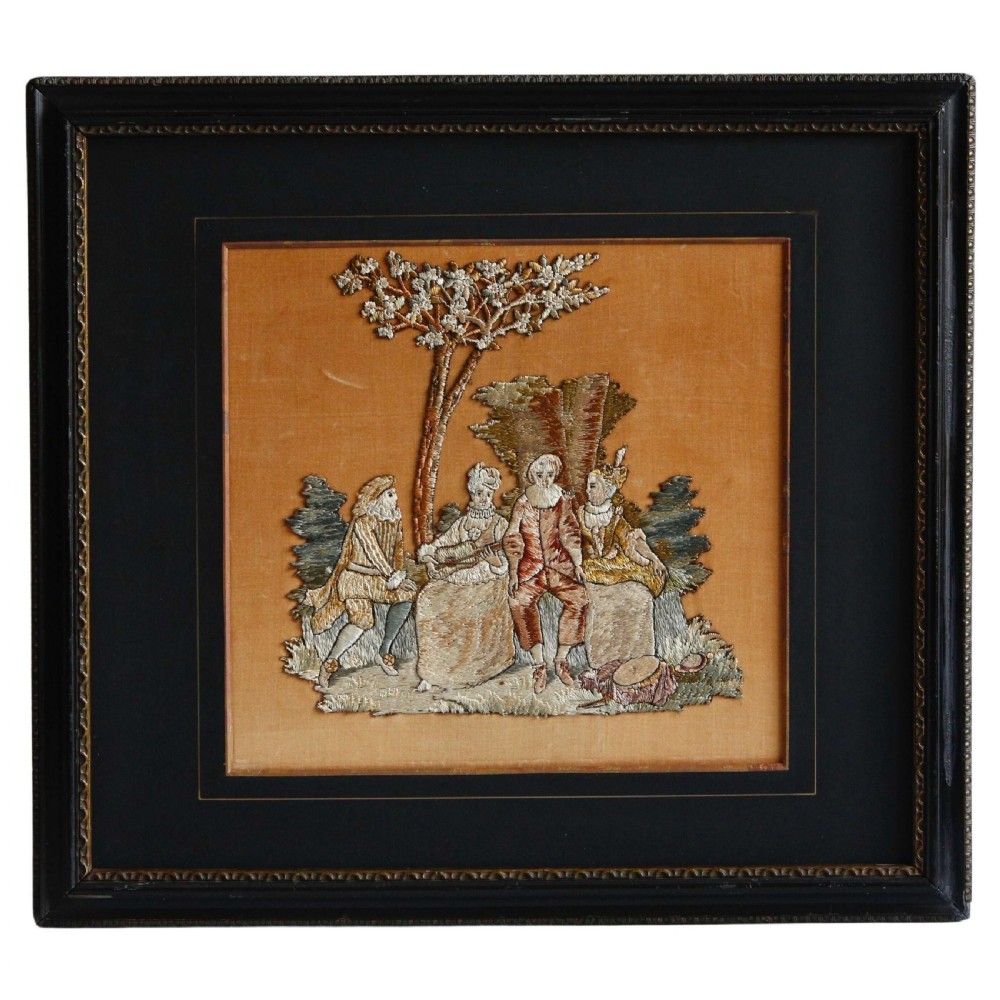 An antique 19th-century framed silk embroidery panel depicting four figures seated outdoors beneath a tree, mounted on an ochre silk background within a black frame.