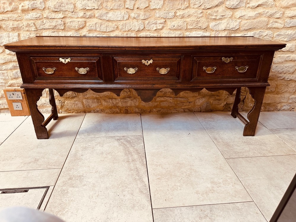 An 18th-century dark oak dresser base featuring three drawers with brass handles and shaped silhouette legs. View X