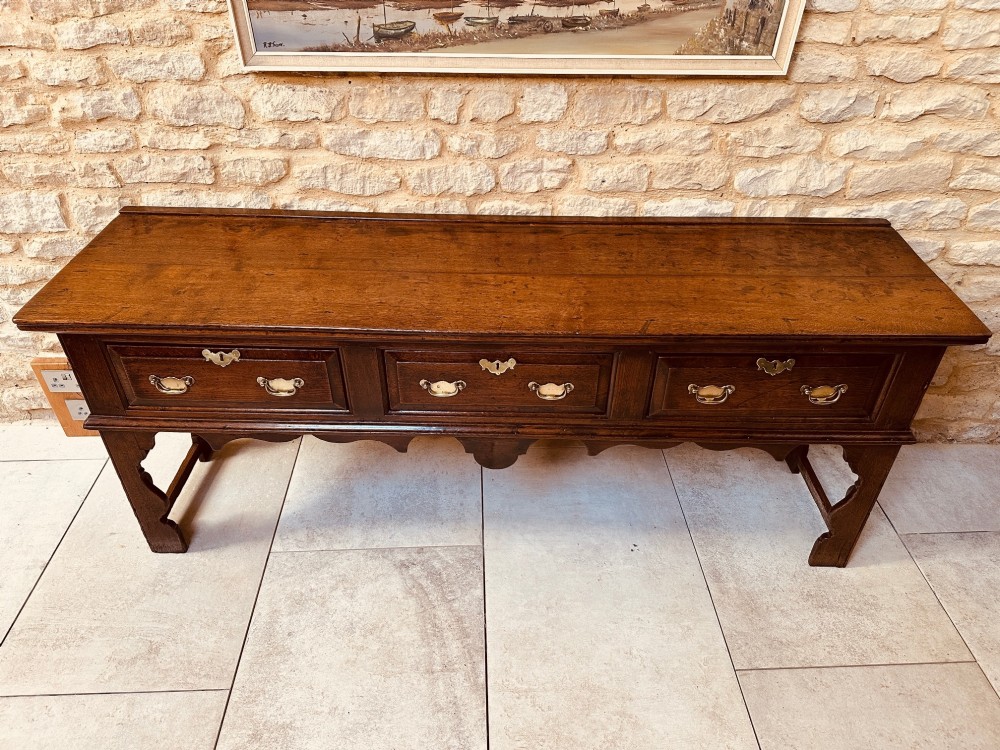 An 18th-century dark oak dresser base featuring three drawers with brass handles and shaped silhouette legs. View XI