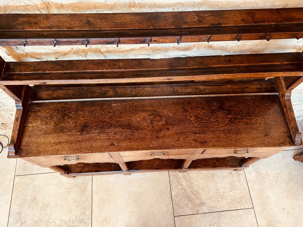 An 18th-century oak potboard dresser featuring an upper open rack with multiple shelves and cup hooks above three frieze drawers and an open lower potboard shelf. View VII