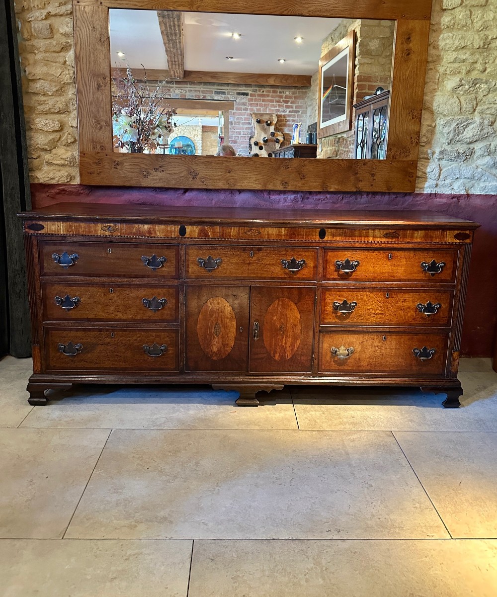 A Georgian oak Lancashire dresser base circa 1800 featuring drawer configuration, central cupboard doors with oval mahogany inlays, and brass handles.