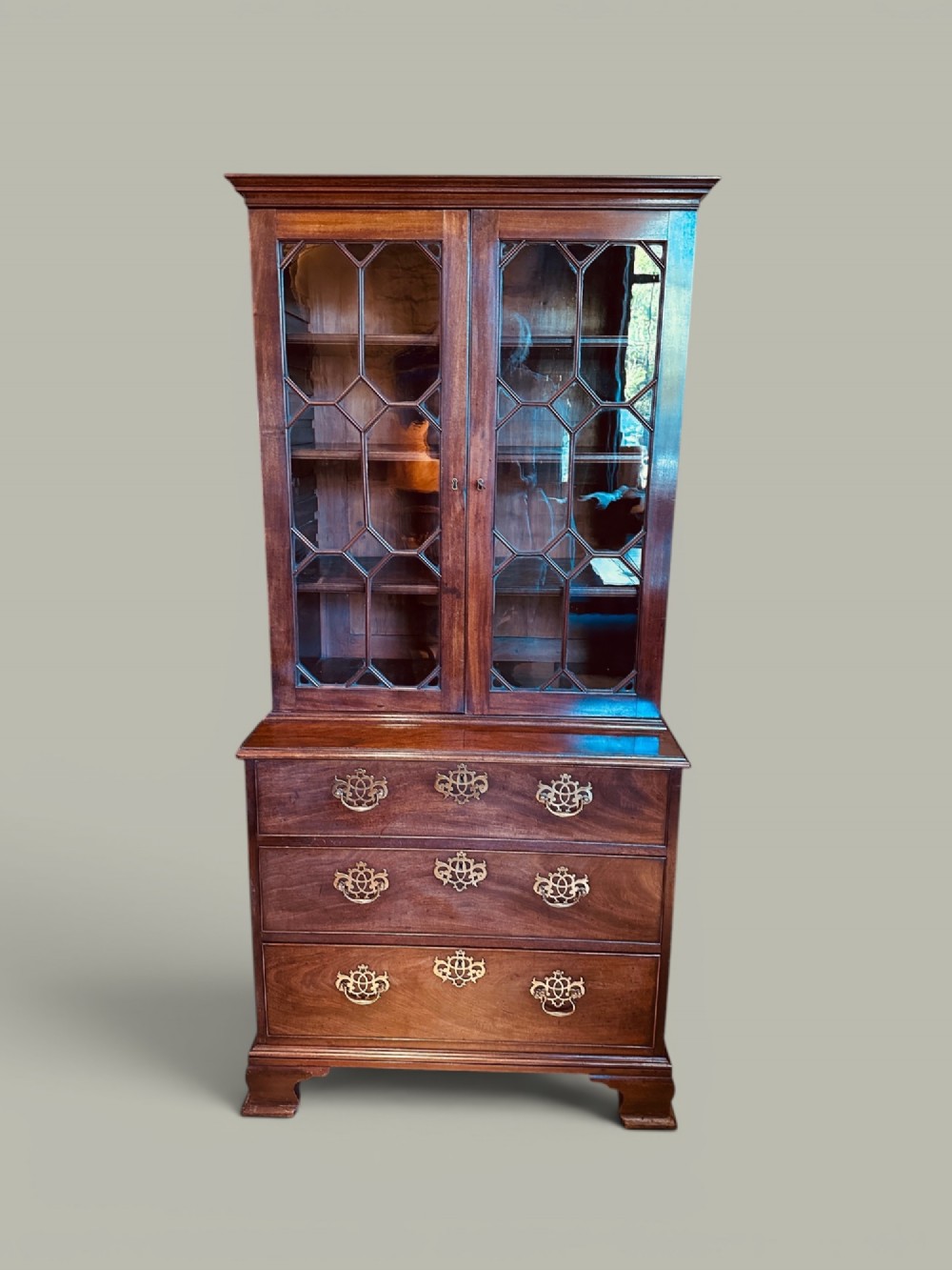An antique mahogany bookcase on chest featuring a two-door astragal glazed upper display section and three long drawers with decorative brass handles below.