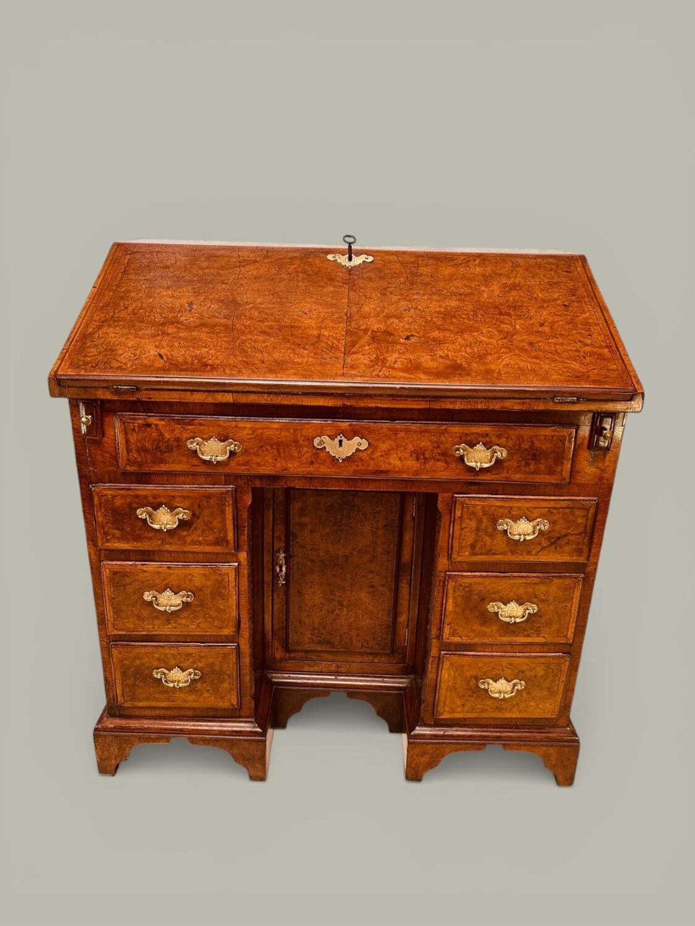 An antique Georgian kneehole bureau in warm brown burr oak featuring multiple small drawers, brass drop handles, a central cupboard, and a hinged top surface.