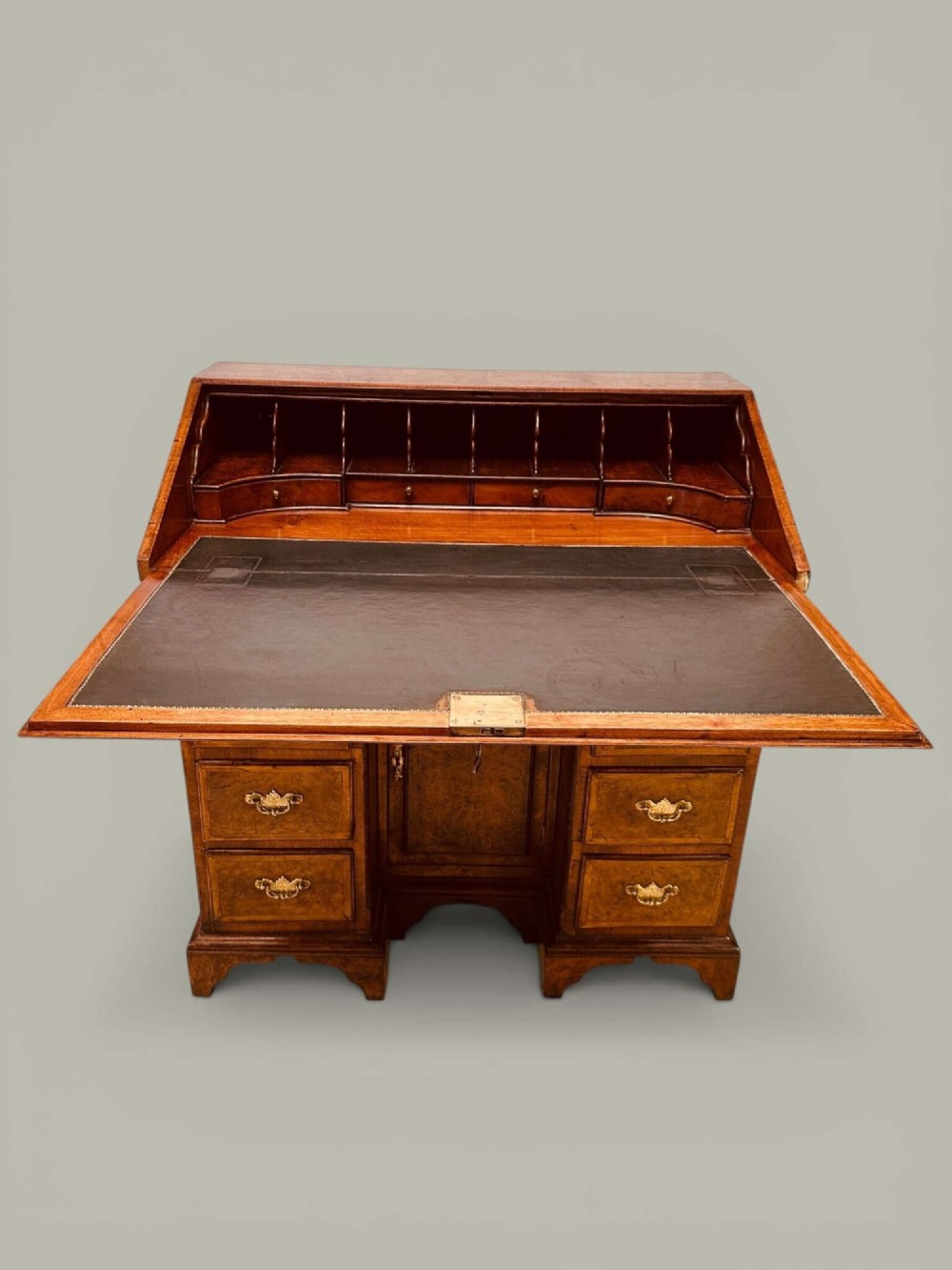 An antique Georgian kneehole bureau in warm brown burr oak featuring multiple small drawers, brass drop handles, a central cupboard, and a hinged top surface. View II