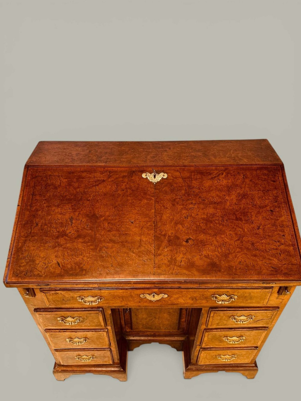 An antique Georgian kneehole bureau in warm brown burr oak featuring multiple small drawers, brass drop handles, a central cupboard, and a hinged top surface. View IV