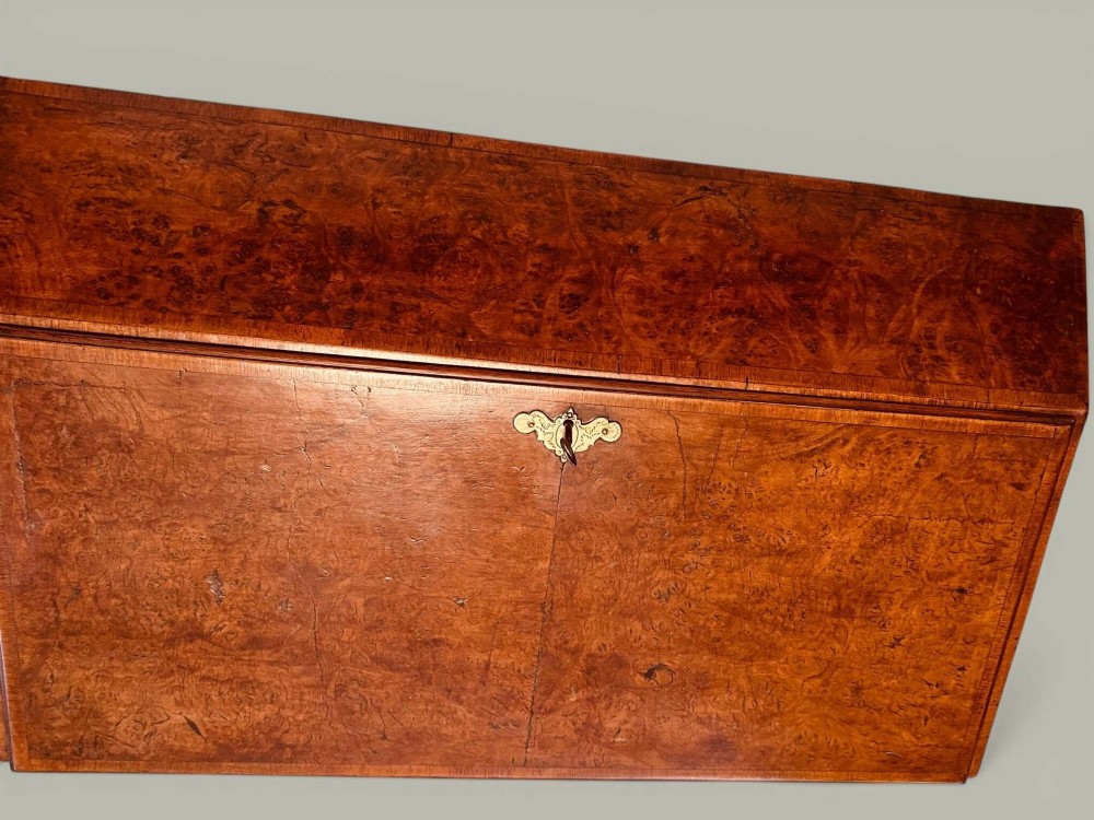 An antique Georgian kneehole bureau in warm brown burr oak featuring multiple small drawers, brass drop handles, a central cupboard, and a hinged top surface. View VI