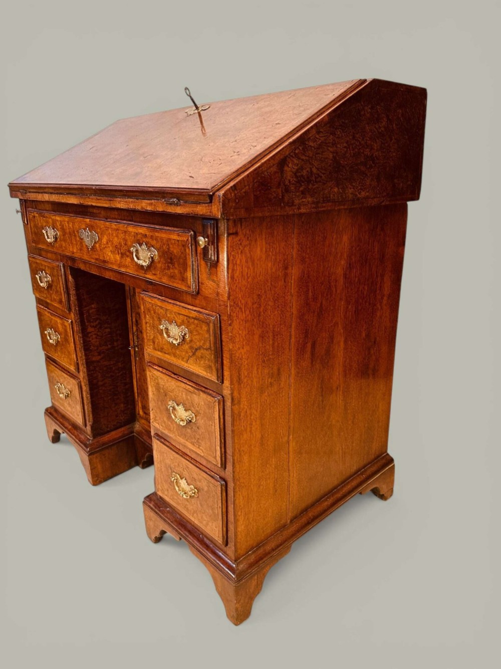 An antique Georgian kneehole bureau in warm brown burr oak featuring multiple small drawers, brass drop handles, a central cupboard, and a hinged top surface. View XII