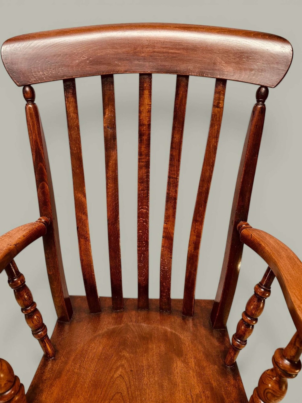 A late nineteenth-century wooden Windsor armchair featuring a curved slat back rest, turned legs, an H-stretcher, and shaped armrests finished in warm brown timber tones. View X