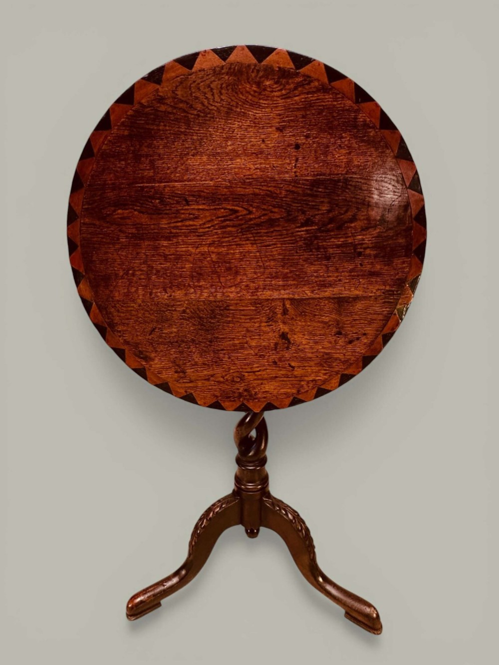 An antique dark oak circular tilt-top pedestal table featuring a geometric inlaid border, a spiral turned column, and carved tripod legs.
