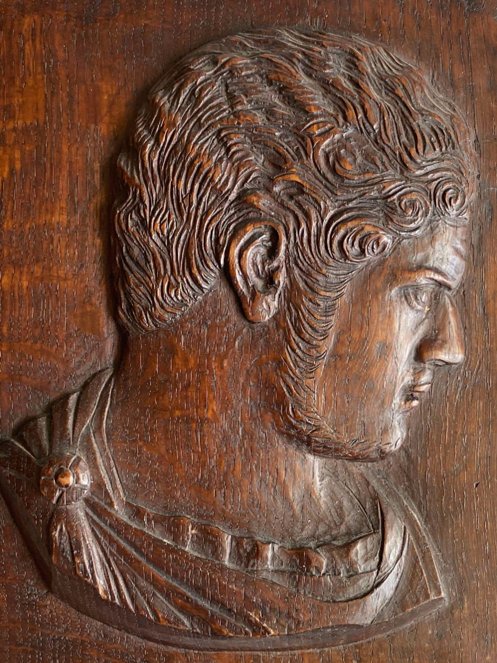 A rectangular dark oak panel featuring a high-relief carving of a Roman emperor profile in profile view. View III