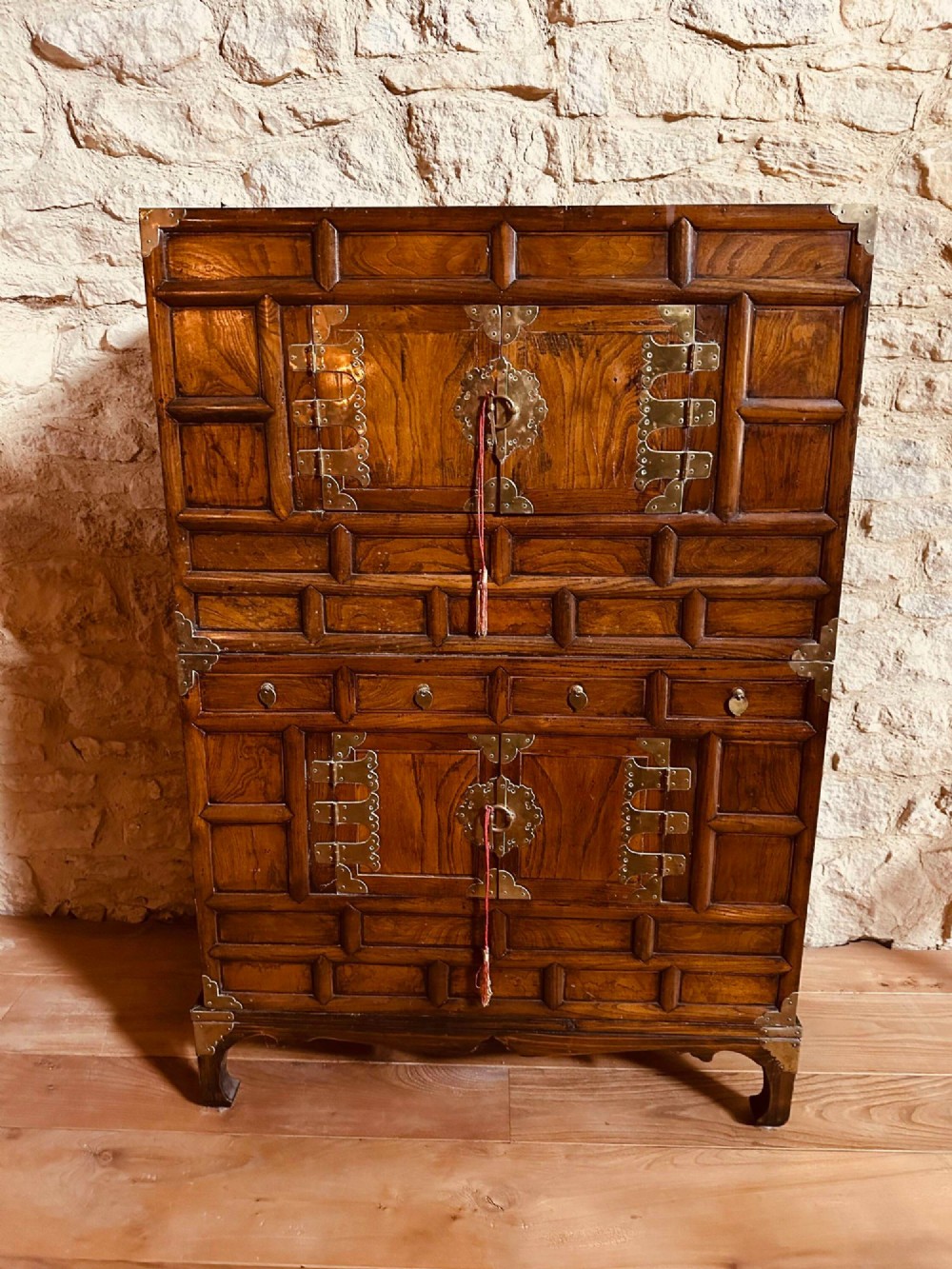 A two-tier Korean elm cabinet with intricate brass corner plates, decorative lock fittings, panelled doors, and red drop tassels.