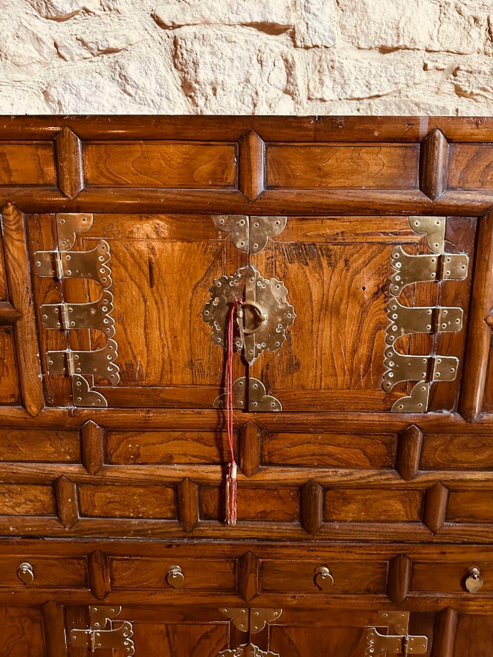 A two-tier Korean elm cabinet with intricate brass corner plates, decorative lock fittings, panelled doors, and red drop tassels. View II