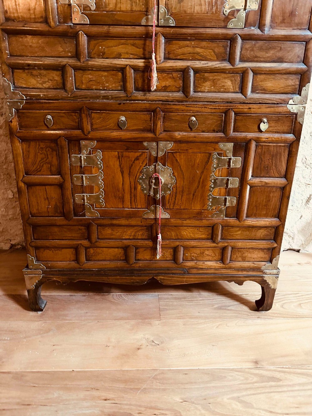 A two-tier Korean elm cabinet with intricate brass corner plates, decorative lock fittings, panelled doors, and red drop tassels. View III