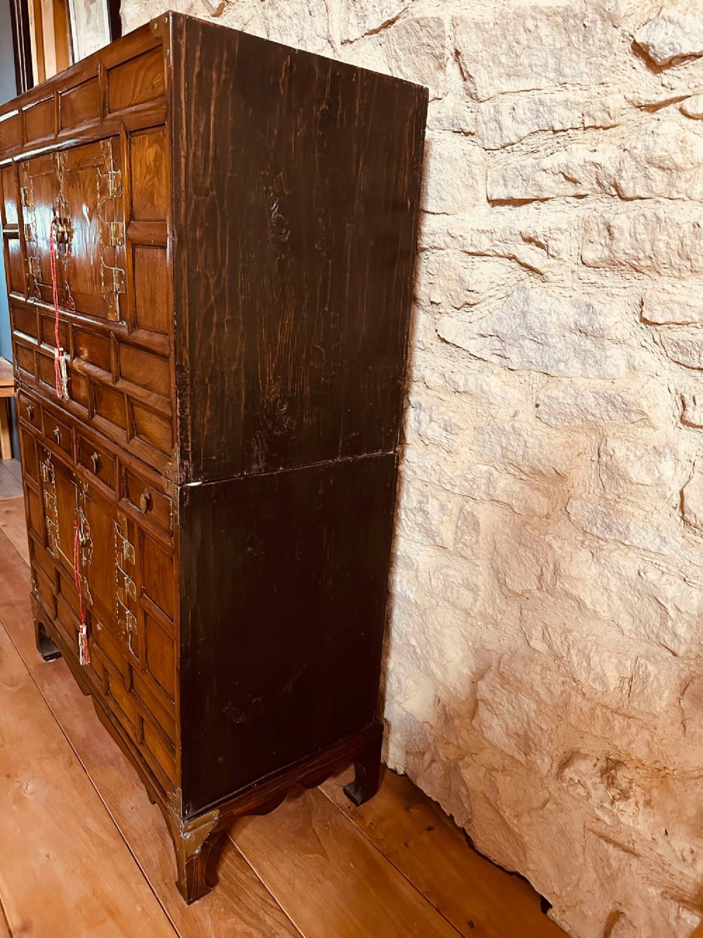 A two-tier Korean elm cabinet with intricate brass corner plates, decorative lock fittings, panelled doors, and red drop tassels. View VII