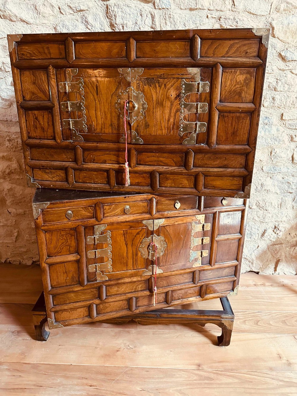 A two-tier Korean elm cabinet with intricate brass corner plates, decorative lock fittings, panelled doors, and red drop tassels. View IX