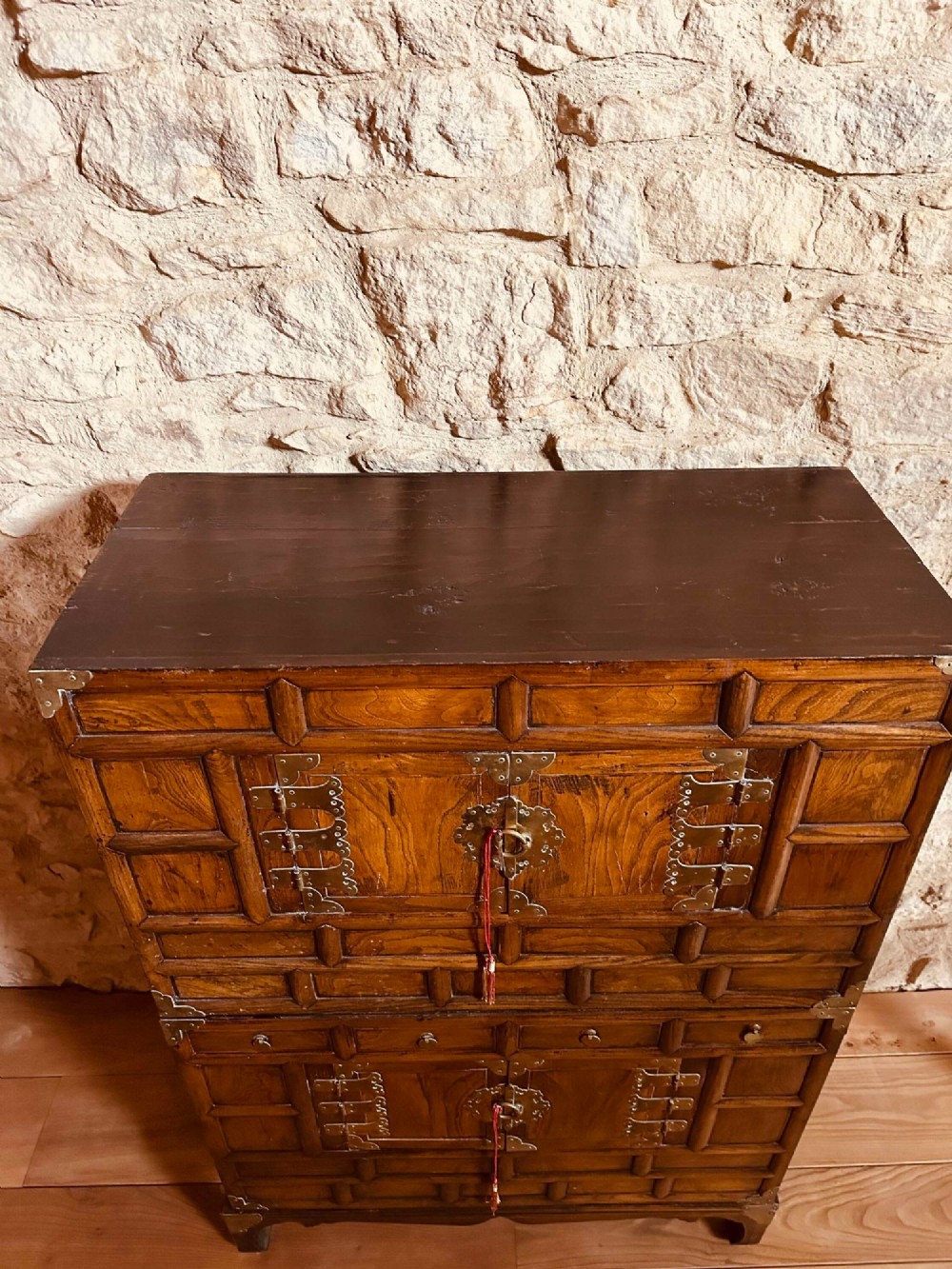 A two-tier Korean elm cabinet with intricate brass corner plates, decorative lock fittings, panelled doors, and red drop tassels. View X