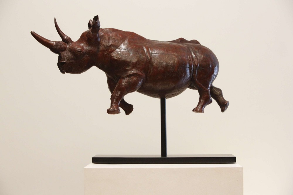 A carved reddish-brown ironwood sculpture of a rhinoceros mounted on a black metal display stand. View II