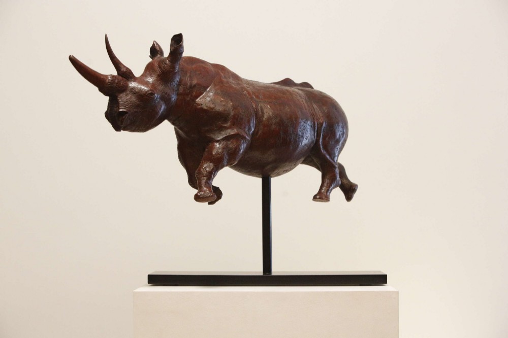 A carved reddish-brown ironwood sculpture of a rhinoceros mounted on a black metal display stand. View XI