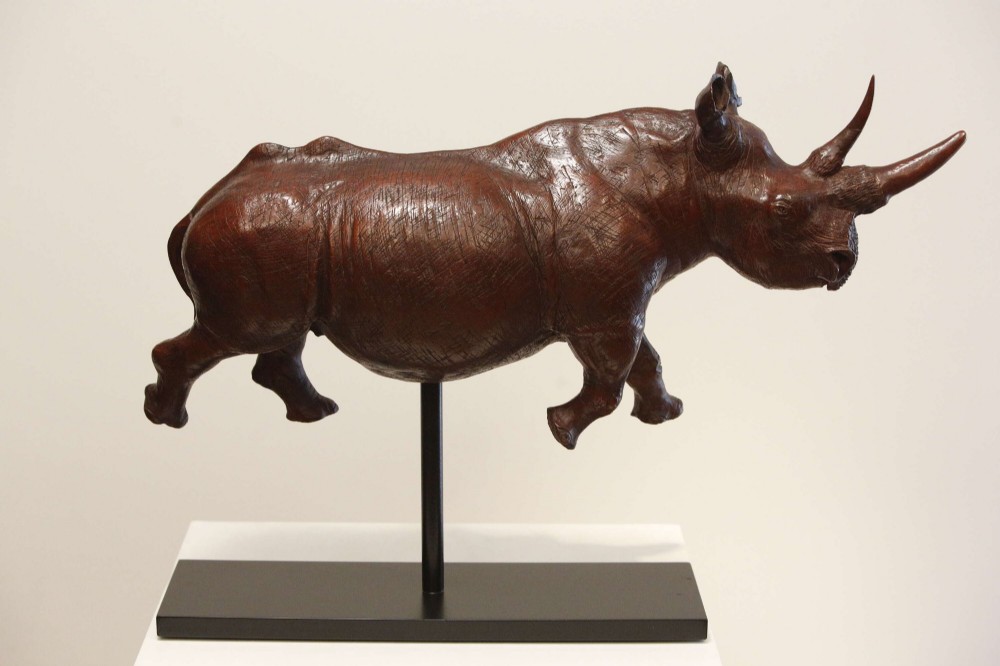 A carved reddish-brown ironwood sculpture of a rhinoceros mounted on a black metal display stand. View XIV