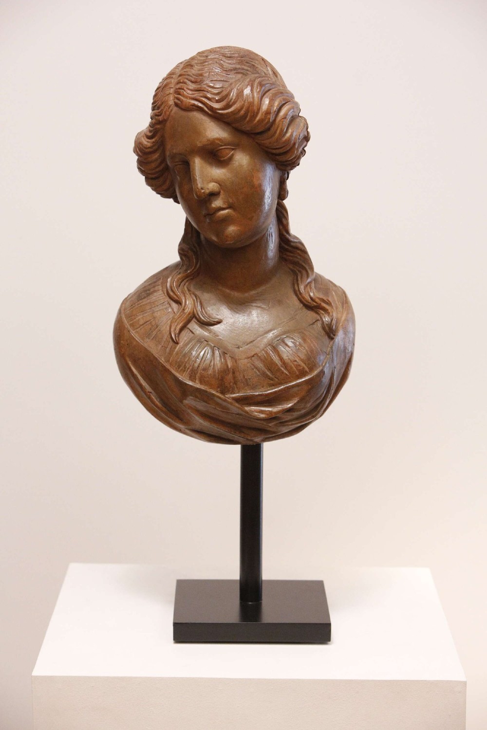 An antique carved wood bust of a maiden with styled wavy hair and draped clothing, mounted on a black metal display stand.