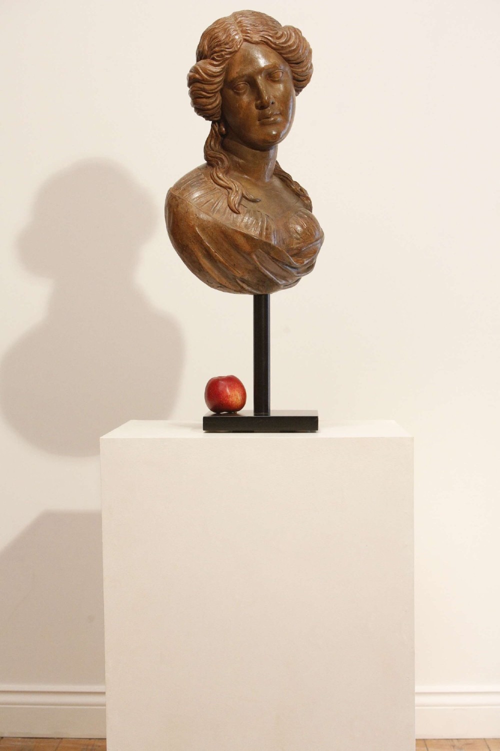 An antique carved wood bust of a maiden with styled wavy hair and draped clothing, mounted on a black metal display stand. View II