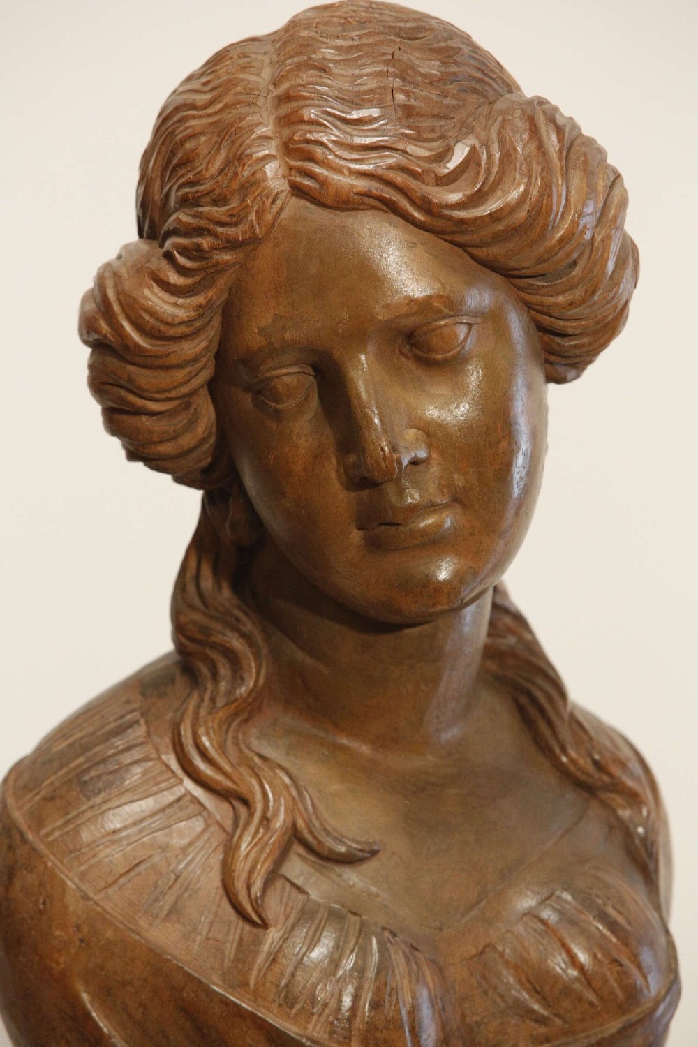 An antique carved wood bust of a maiden with styled wavy hair and draped clothing, mounted on a black metal display stand. View III