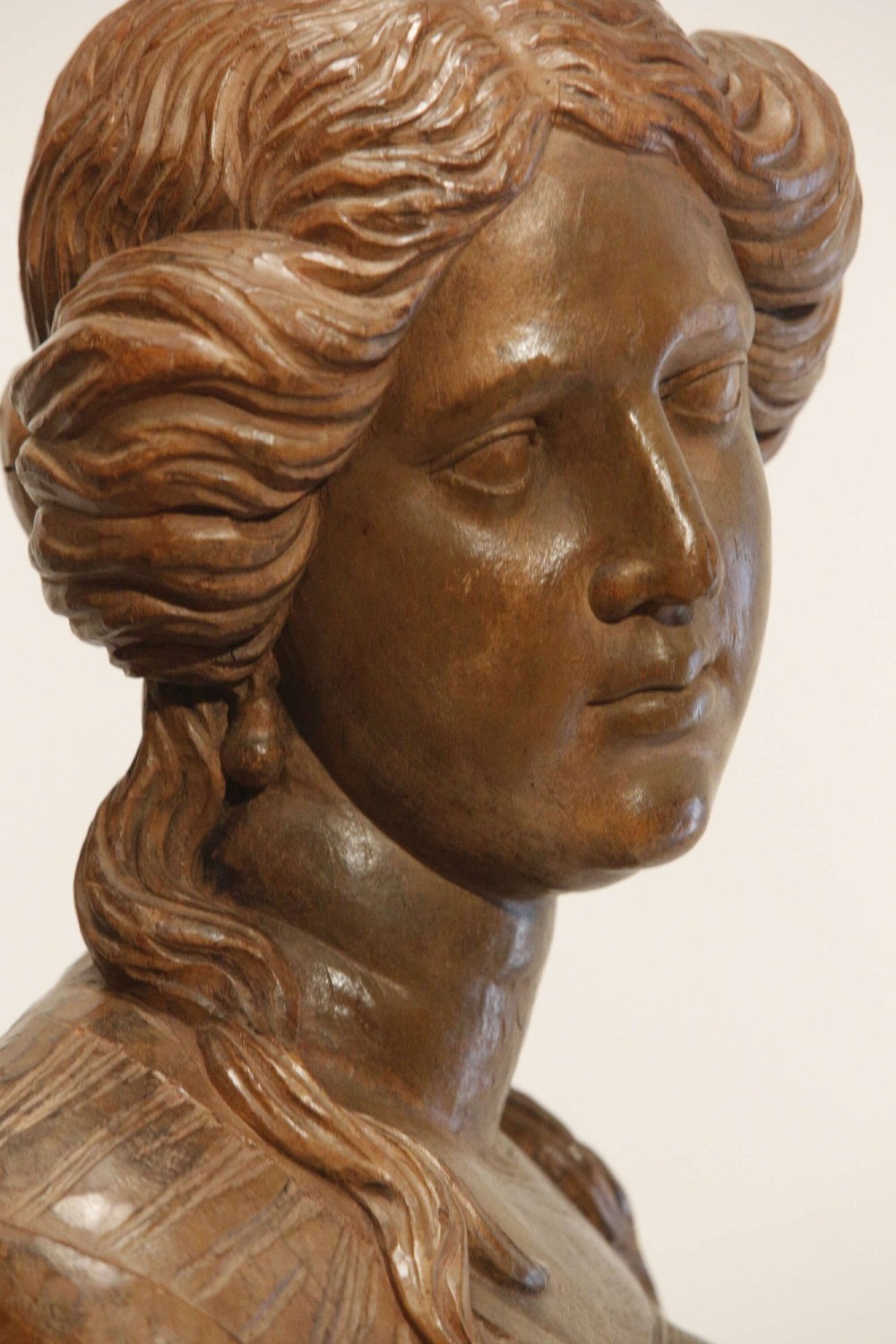 An antique carved wood bust of a maiden with styled wavy hair and draped clothing, mounted on a black metal display stand. View IV