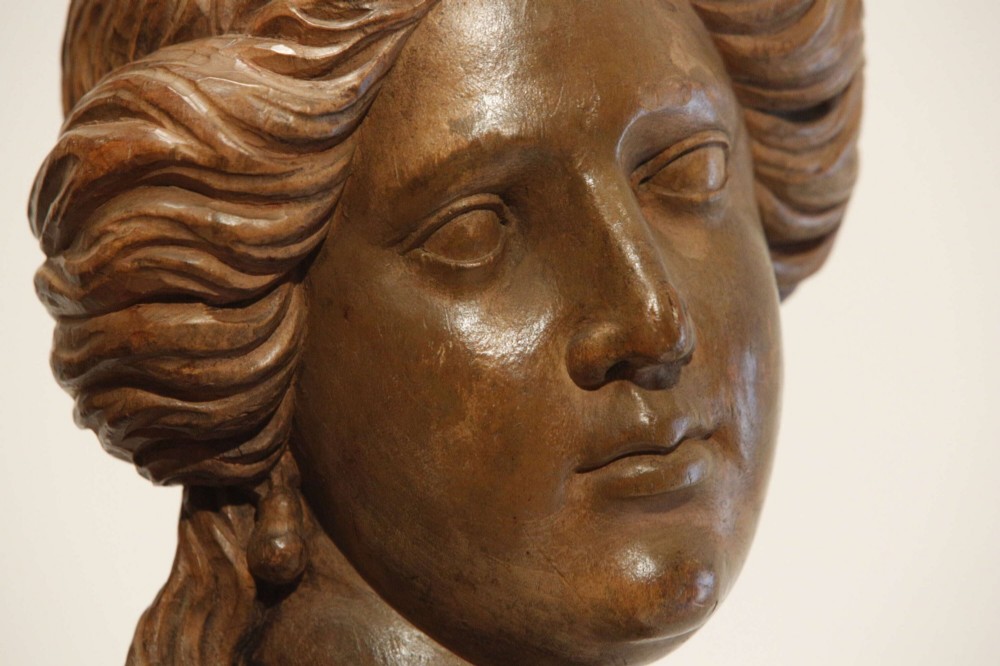 An antique carved wood bust of a maiden with styled wavy hair and draped clothing, mounted on a black metal display stand. View V