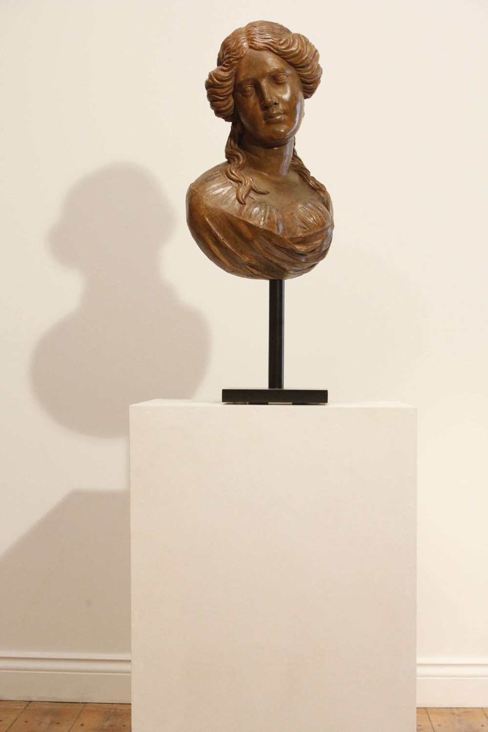 An antique carved wood bust of a maiden with styled wavy hair and draped clothing, mounted on a black metal display stand. View VI