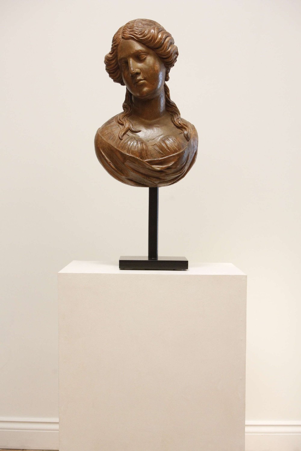 An antique carved wood bust of a maiden with styled wavy hair and draped clothing, mounted on a black metal display stand. View VII