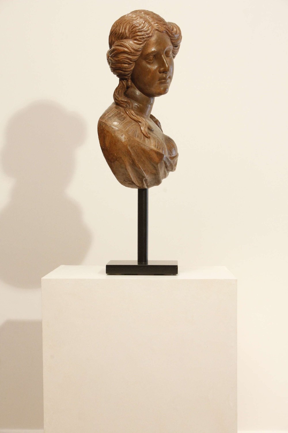An antique carved wood bust of a maiden with styled wavy hair and draped clothing, mounted on a black metal display stand. View VIII