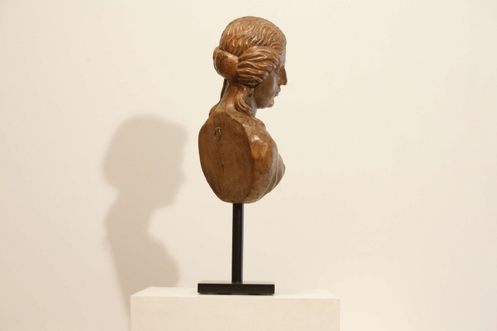 An antique carved wood bust of a maiden with styled wavy hair and draped clothing, mounted on a black metal display stand. View IX