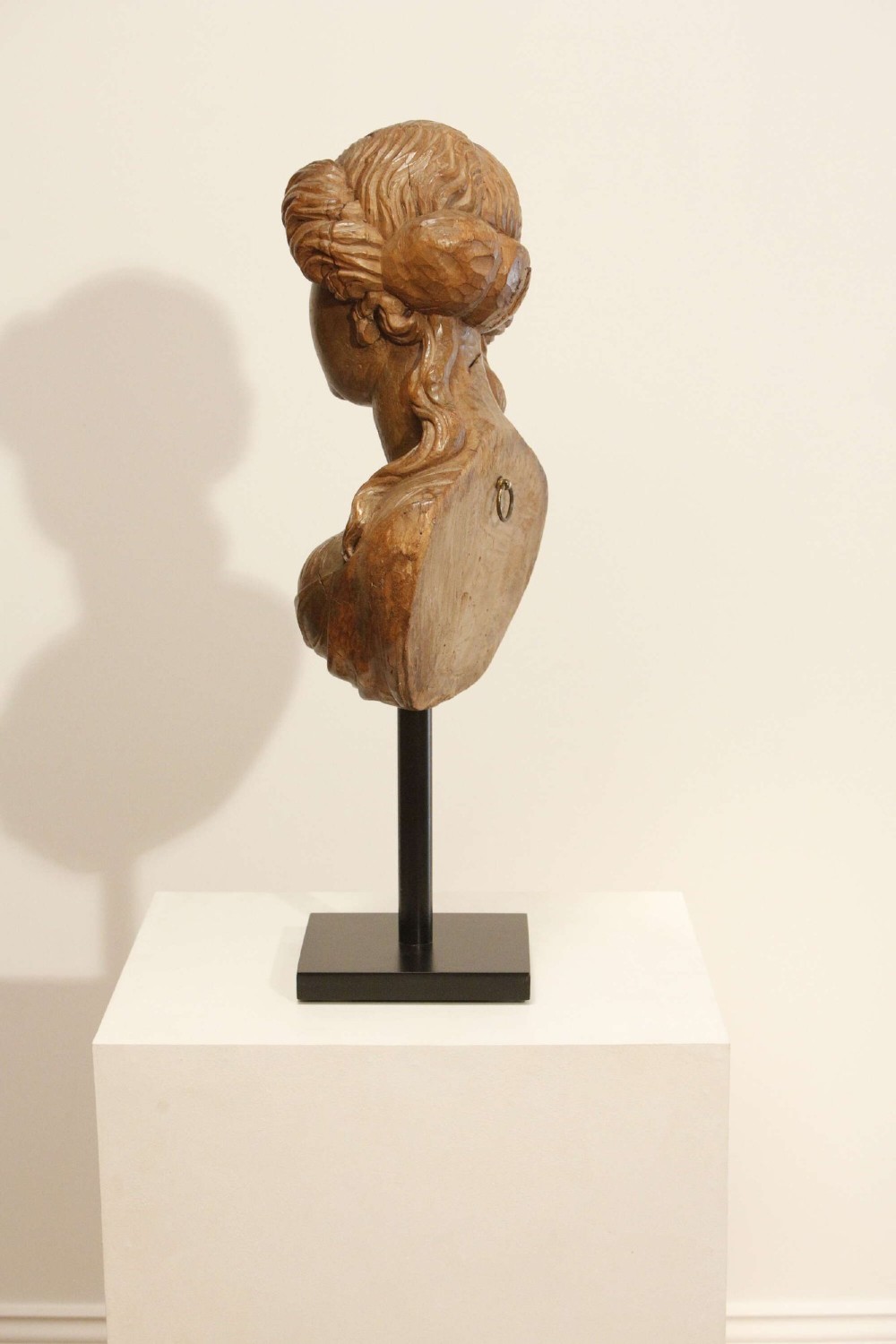 An antique carved wood bust of a maiden with styled wavy hair and draped clothing, mounted on a black metal display stand. View X